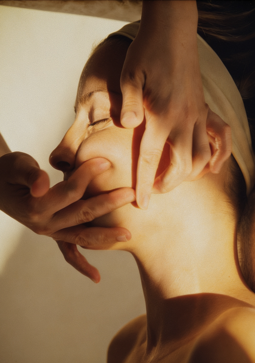A close-up lifestyle shot of a woman receiving a facial massage, with her face partially covered by her hands and another hand gently holding her cheek. She is wearing a light beige headband, and the scene is illuminated by warm, dramatic side lighting.